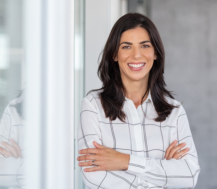 Portrait of middle aged businesswoman in modern office looking at camera. Confident business woman with arms crossed standing while leaning against glass wall. Proud brunette woman smiling in formalwear with copy space.